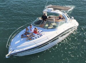 A sleek white motorboat is anchored on calm water. Two people are lounging on striped blue and white sun pads at the front of the boat; one is wearing a red top and the other is in a white shirt. A third person is seated at the helm, wearing a black shirt and a cap. In the background, another individual is visible, enjoying a drink. The boat features a modern design with a spacious cockpit and a sunshade. The scene conveys a relaxed, leisurely atmosphere on the water.