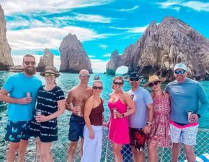 A group of eight people stands on a boat with a scenic backdrop of rocky cliffs and turquoise water. They are dressed in casual, summery attire, with some wearing sunglasses and hats. The individuals are holding drinks in metal cups and are smiling, enjoying their time together. In the background, notable rock formations rise from the water, under a bright blue sky with scattered clouds.
