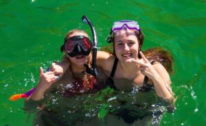 Two individuals are enjoying a snorkeling experience in clear, green water. One person is wearing a black snorkel mask and has a snorkel attached, while the other has a purple snorkel mask. They are both smiling and posing for the photo, with one making a peace sign and the other showing a "shaka" hand gesture. Sunlight reflects off the water, creating a vibrant atmosphere.