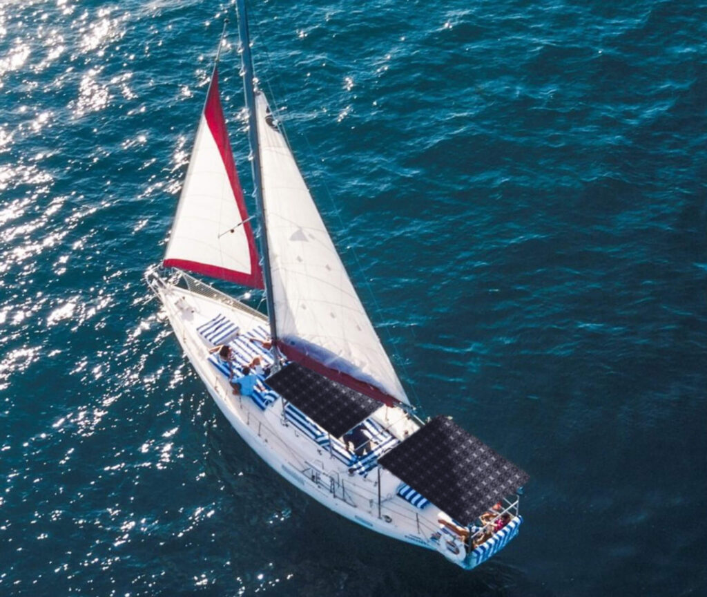 A sailboat is depicted from an aerial perspective, gliding over shimmering blue waters. The boat features a white hull with red accents on the sails. Solar panels are installed on the deck, and there are striped blue and white cushions visible, suggesting a comfortable seating area. The sunlight reflects off the water, creating a sparkling effect around the vessel.